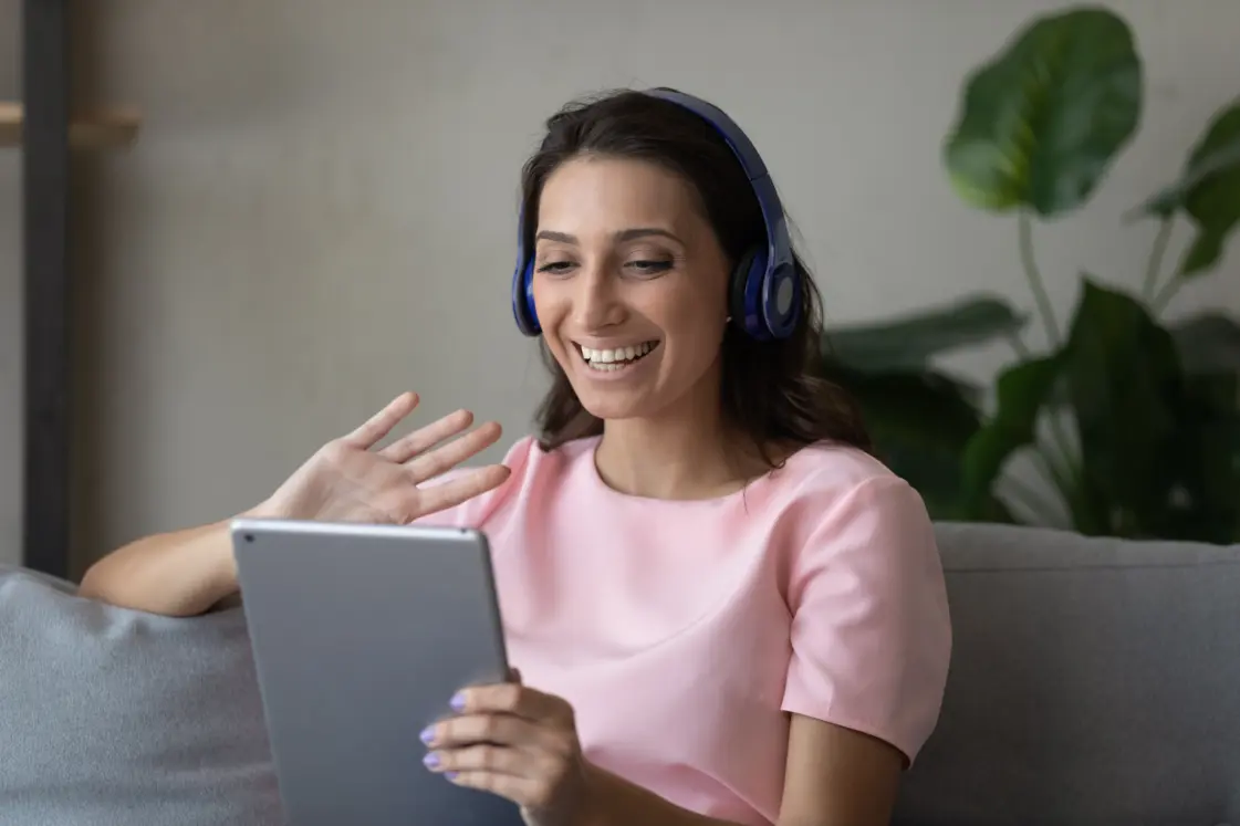 Lady is waving to her tablet with headphones on, during an online therapy session for anxiety.